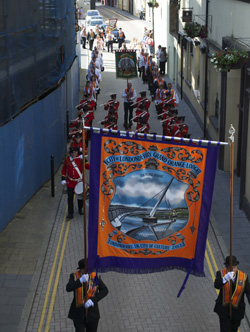 The Orange Order, a Unionist organization, marches in preparation for a large march on the 12th of July. The banner for the City of Londonderry lodge depicts the Peace Bridge, a pedestrian bridge built to connect the ‘Waterside’ and ‘Cityside’ areas of the city. 