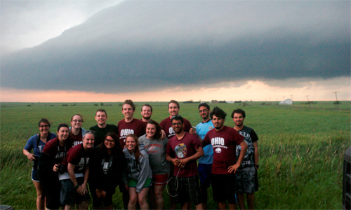 Dr. Jana Houser led 12 undergraduates and two graduates on a storm-chasing trek through the Central Plains. Shown here standing in a field as a group with storm clouds behind them.