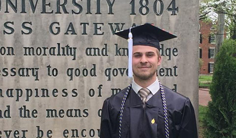 Justin Bergh in cap and gown in front of College Green gate sign
