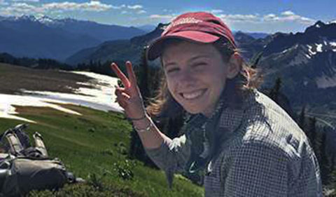 Jocelyn Pettit smiling with right hand held up with fingers in V shape, while sitting on mountain top with spots of snow in Mount Rainier National Park in 2016