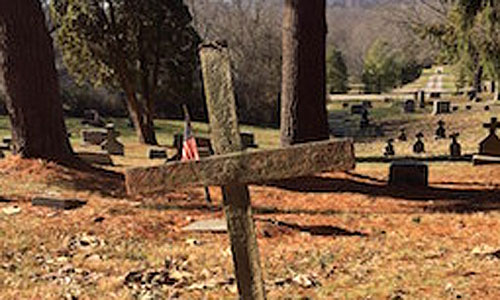 OHIO sophomore Alison Harper studied three cemeteries, including St. Patrick’s (pictured here), to find demographic and mortality information about a rural Appalachia population. Her work is supported by the Provost’s Undergraduate Research Fund. Photographer: Kelee Riesbeck This photo shows a wooden cross in the foreground, with stone markets in the background.