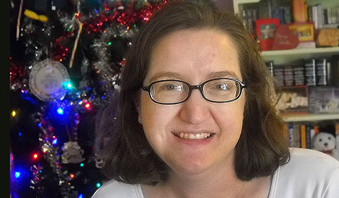 Jenny Shank, smiling, with decorated Christmas tree and bookshelves behind her
