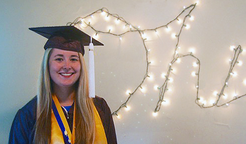 smiling Rachel Neal in cap and gown with honor cord and honor sash standing in front of light string that is shaped on the wall behind her to spell Ohio