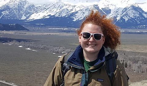 A smiling Adrianne Talmadge, wearing sunglasses and with wind-blowing hair and Tetons in the background