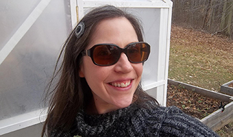 A smiling Lauren Ketcham in sunglasses and sweater, standing in front of white wood framed greenhouse with plastic walls