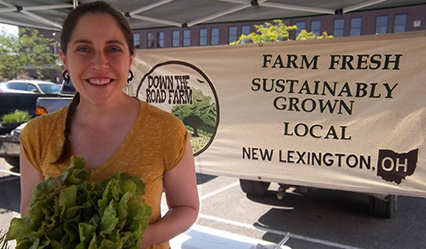 A smiling Lauren Ketcham holding a large head of leaf lettuce with Down the Road Farm banner behind her in a farmers market tent