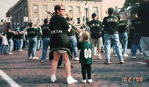 Alumni band members with instruments as they make their way down Court Street in Athens Ohio during the 2010 reunion. Stacie has turned back to look behind her as she stands with her trombone at the ready with young daughter at her side.