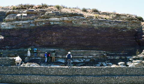 Ichnologists investigate a large Triassic burrow. Photo taken from a distance, with scientists working on a ledge.