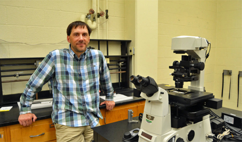 Anthony Stender in his microscopy lab at Clippinger. He is shown here leaning against a work bench with a microscope on a table in front of him.