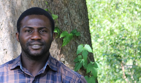 portrait of a dark skinned man standing in front of a tree trunk surrounded by bright green leaves