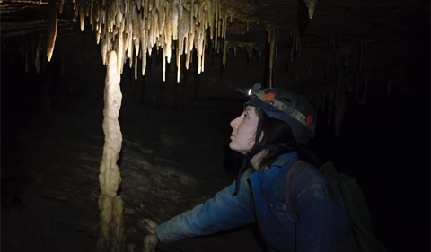 a white female with dark hair dressed in a blue caving suite and helmet. A helmet light is shining on a stalactite in an otherwise dark cave