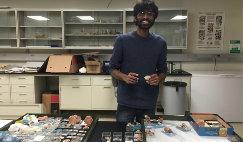 a snapshot of Ranjeev Epa, a man with dark skin, dark hair, a dark full beard wearing glasses and who is wearing a dark blue sweater and blue jeans stands behind a collection of small fossils displayed on a table in front of him. The background is a research lab with cream cabinets and glass shelves on the wall.
