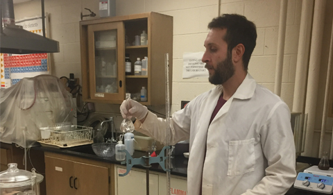 Lucas Howard holds a glass flask containing a water sample as he prepares it for analysis. He is wearing a long white lab coat, latex gloves, and is surrounded by the detrious of a geochemistry research lab.
