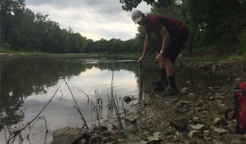 Lucas Howard dips a water collection instrument into the Maumee River as he balances between the bank and a jutting rock. He is wearing dark shorts, a red tshirt, and a gray ballcap. You can see the banks of the river in the background surrounded by trees.