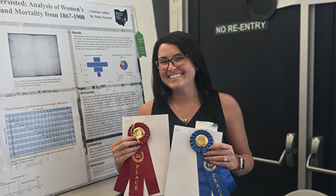 A smiling Courtnee Ambos holding two award ribbons while standing in front of research poster