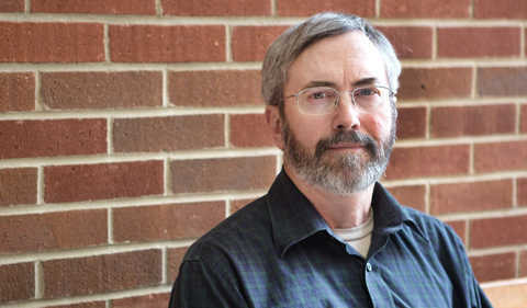 a portrait of a man with glasses, graying hair and full beard wearing a blue button down shirt standing in front of a brick wall